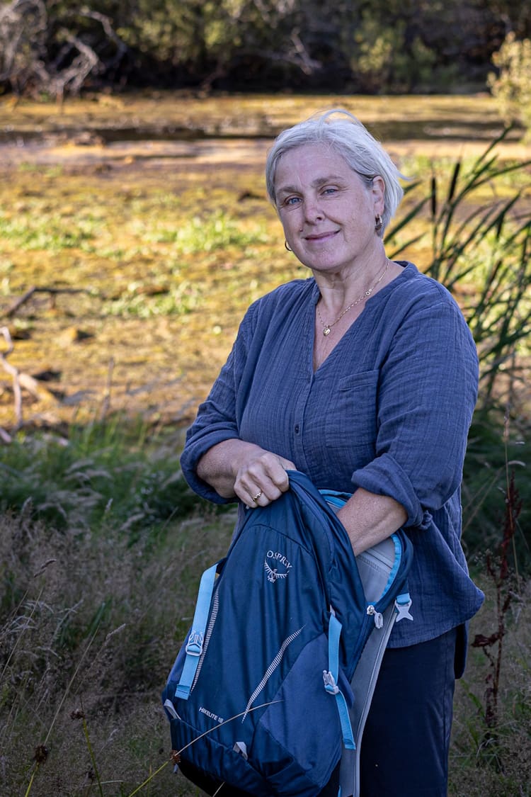 En smilende Anja Vindal står i naturen med sin rygsæk i hånden og kigger smilende på dig. Hun tilbyder naturterapi